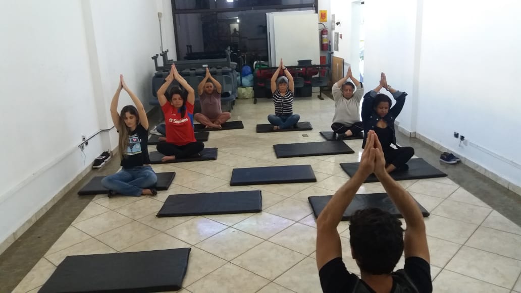 Imagem colorida, com diversas pessoas em uma posição de yoga, sentada em tapetes.